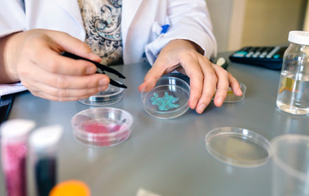 Unrecognizable female chemist technician hands holding a blue glitter sample over petri dish on a...