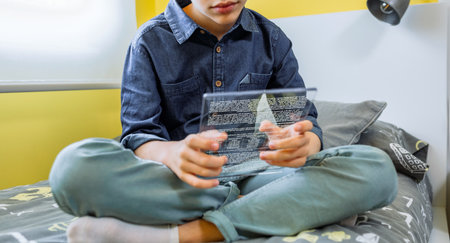 Unrecognizable boy using futuristic transparent tablet to read ebook sitting over his bed. Digital display with glass screen concept. Relaxed young kid holding portable device to study at home.の写真素材