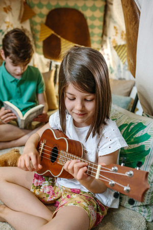 Portrait of smiling little girl playing ukulele while boy reading book on handmade teepee. Children having fun in cozy diy shelter tent in their house. Vacation camping at home or staycation concept.の写真素材