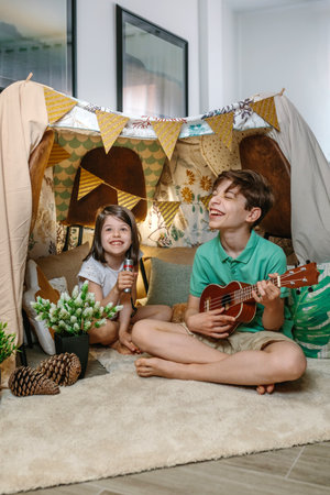 Happy children playing a ukulele and singing on handmade shelter tent in living room. Boy and girl...