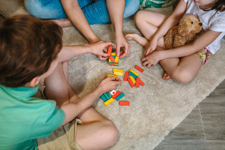 Unrecognizable mother and sons having fun playing wooden stacking piece game over carpet at home. Single parent family enjoying together in leisure time. Happy little girl holding stuffed teddy dog.の写真素材