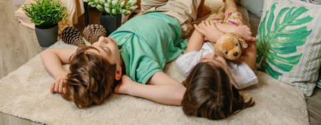 Banner of serene boy and little girl embracing stuffed dog resting surrounded by cushions and plants at home. Two children relaxing lying over rug in a warm and playful ambiance at home.の写真素材