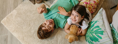 Banner of children laughing and resting lying over rug in cozy ambiance at home. Happy boy pointing with finger up and smiling little girl looking while embracing stuffed dog surrounded by cushions.の写真素材