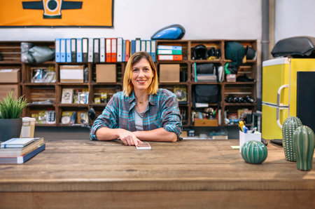 Portrait of smiling blonde young female employee looking at camera behind of industrial shop...