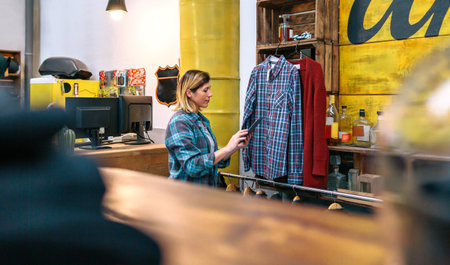 Blonde woman looking digital tablet while checking stock on second hand clothing store. Young female working on store with industrial style. Sustainable shopping and circular apparel economy concept.の写真素材