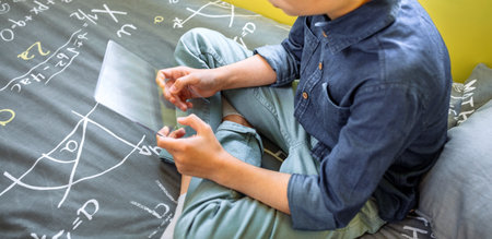 Unrecognizable young kid holding digital display with glass screen sitting over his bed. Futuristic...