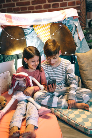 Smiling children looking mobile phone together while chatting on social media application sitting on shelter tent at home. Happy little girl embracing rag doll next to boy using cell in a cozy teepee.の写真素材