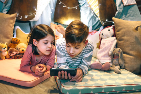 Children watching content on cellphone without parental supervision at home. Girl and boy lying together while chatting with strangers on social network app. Screen addiction dangers concept.の写真素材