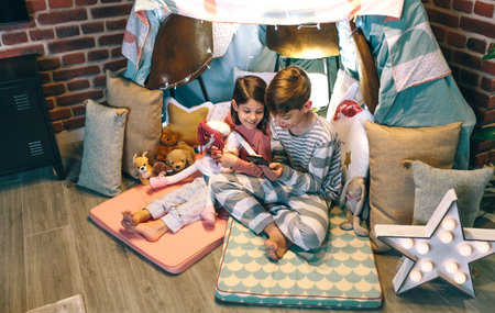 Smiling children looking mobile phone together while chatting on social media application sitting on shelter tent at home. Happy little girl embracing rag doll next to boy using cell in a cozy teepee.の写真素材