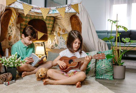 Little girl playing ukulele while boy reading book on handmade teepee in living room. Children having fun in a cozy diy shelter tent in their house. Vacation camping at home or staycation concept.の写真素材