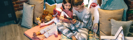 Banner of smiling children looking mobile phone together while chatting on social media application sitting on tent at home. Happy little girl embracing rag doll next to boy using cell in cozy teepee.の写真素材