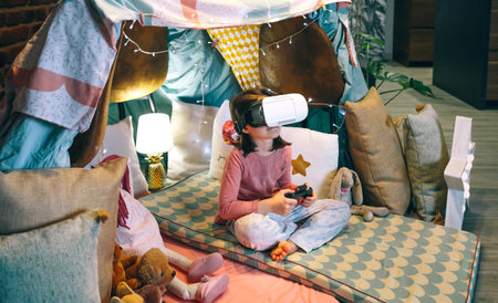 Digital native child watching metaverse world through VR glasses sitting over mats in tent. Girl wearing virtual reality headset and playing games in teepee. Cyberspace and tech recreation concept.の写真素材