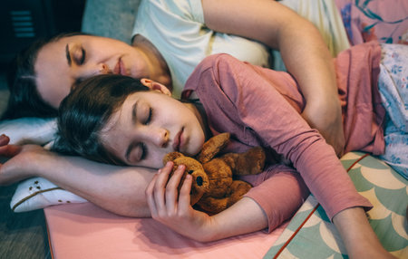Portrait of serene daughter embracing a teddy bear lying next to mom over mats at night. Close up of woman sleeping and hugging little girl with closed eyes. Security and family protection concept.の写真素材