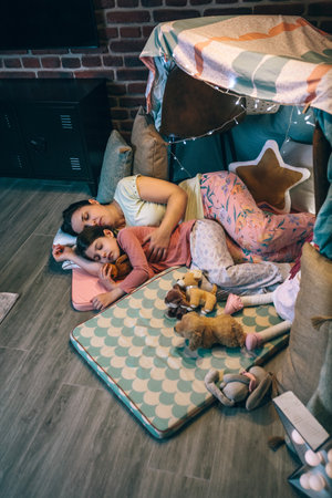 Loving mother and daughter sleeping together lying over mats in cosy diy tent. Woman embracing to little girl asleep surrounded by cushions in handmade teepee at night. Serene and peaceful atmosphere.の写真素材