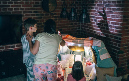 Back view of mother and two children having fun playing with shadow puppets over brick wall at home pajama party. Woman making rabbit silhouette with her hands in shade theatre in living room.の写真素材