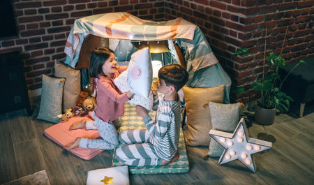 Happy siblings having fun in pillow fight on night party at living room. Cheerful boy and girl with pajama laughing and playing in cushion battle at home in front of tent. Domestic family life conceptの写真素材