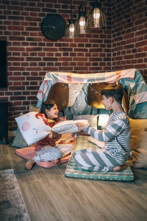 Happy children with pajama having fun in a pillow fight on handmade shelter tent in living room. Excited boy and girl laughing and playing in cushion battle on teepee. Entertainment at home conceptの写真素材