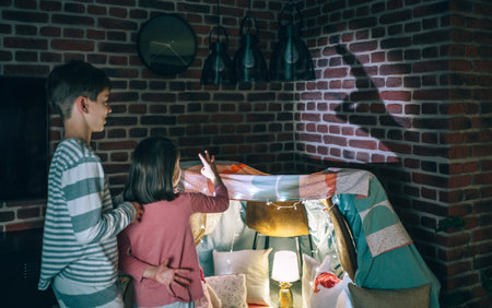 Two children in pajama having fun playing with shadow puppets over brick wall at home. Back view of sister making rabbit silhouette with hands in shade game next to her brother in living room at nightの写真素材