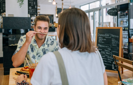 Young man playing with a chopstick like it is a mustache while enjoying meal with his girlfriend at a cozy cafe, sharing laughter and love over their lunch date. Happy couple having fun in restaurant.の写真素材