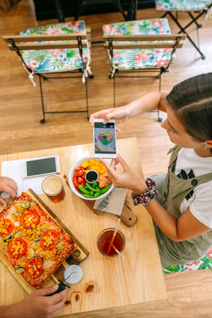 Top view of happy young woman taking photo of delicious fresh poke bowl salad while enjoying lunch with her friend at a cozy cafe. Healthy lifestyle and vegan food concept.の写真素材