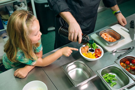 Professional chef pouring soy sauce on colorful poke bowl with salmon, vegetables and mango fruit, while little girl pointing her finger at it, in restaurant kitchen. Healthy eating education concept.の写真素材
