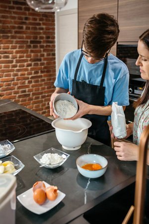Teenage boy wearing an apron pours flour into a mixing bowl while baking with his mother in a modern kitchen, surrounded by various ingredientsの写真素材