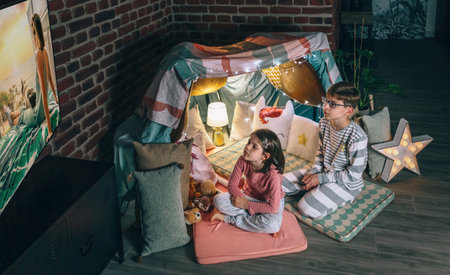 Boy and girl looking tv show sitting over mats on living room at night. Two concentrated children in front of handmade teepee resting while watching movie on television at home. Domestic life concept.の写真素材