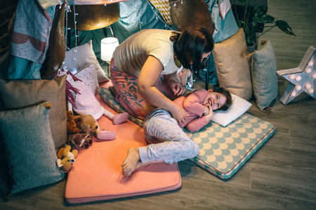 Top view of young daughter laughing while mother tickling in a diy tent camping. Woman and girl playing and having fun over cozy mats in night pajama party. Weekend activities at home concept.の写真素材