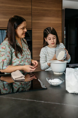 Young daughter pours flour into a mixing bowl while baking with his mother in a modern kitchen,...
