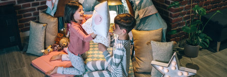 Banner of happy siblings having fun in pillow fight on night party at living room. Cheerful boy and girl with pajama laughing and playing in cushion battle at home. Domestic family life conceptの写真素材