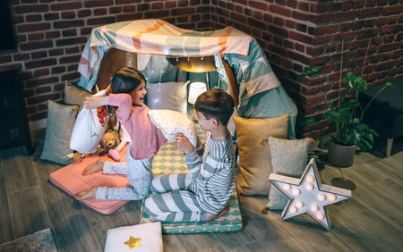 Excited boy and girl laughing and playing in cushion battle on teepee. Happy children with pajama having fun in a pillow fight on handmade shelter tent in living room. Entertainment at home conceptの写真素材