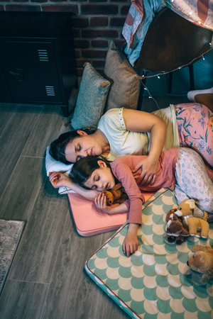 Mother embracing to little daughter little asleep surrounded by cushions at night. Loving woman and girl sleeping together lying over mats in cosy diy tent. Security and family protection concept.の写真素材