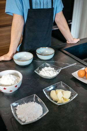 Unrecognizable young pastry chef is arranging bowls of flour, sugar, butter, and eggs on a sleek,...