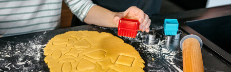 Banner of unrecognizable young child using cookie cutters of various shapes on a rolled out dough on...