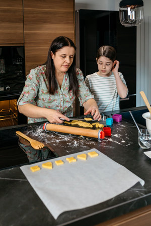 Mother arranging shapes of unbaked cookies on oven paper sheet for baking with her daughter in modern kitchen, sharing warm moment of culinary learning and family connection. Copy space in lower area.の写真素材