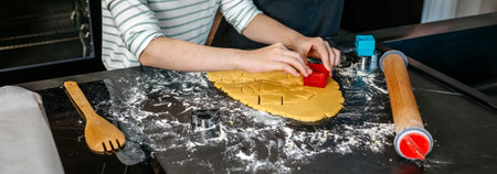 Unrecognizable young girl cutting cookies from dough with various cookie cutters while boy helping to her in a modern kitchen. Brother and sister making festive dessert together for a party at homeの写真素材