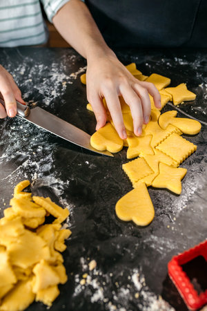 Unrecognizable young girl pastry chef cutting heart shaped cookies from rolled dough with knife on black kitchen counter, with dough scraps and cookie cutters visible. Copy space available on image.の写真素材