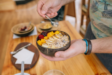 Close up of unrecognizable chef adding seeds with a small spoon to healthy breakfast bowl made with mango, banana, raspberries, cereals and muesli during a cooking master class. Vegan meal concept.の写真素材