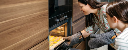 Banner of mom and her daughter are taking a baking pan full of golden cookies out of the oven in...
