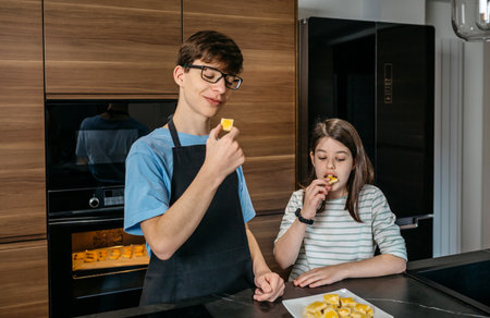 Happy teenage brother wearing apron and sister are tasting freshly baked cookies in a modern kitchen, enjoying the fruits of their labor. Two children eating delicious biscuits baking by them.の写真素材