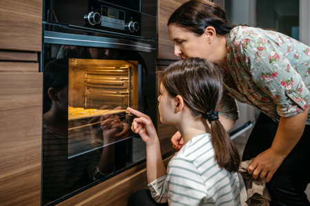 Mother and daughter are carefully watching cookies baking in the oven, anticipating the delicious outcome of their work. Young girl pointing with her finger to biscuits. Family time cooking concept.の写真素材