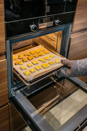 Close up of unrecognizable hand wearing oven mitts taking a baking pan full of golden cookies out of the oven. Home safety concept in the kitchen.の写真素材