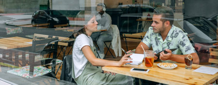 Banner of two young students sitting at a wooden table while discussing their studies and learning together, with drinks and snacks on the table, seen through the cafe glass.の写真素材