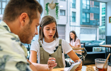 Portrait of young girl and her male university classmate are studying together at a table in a cafe, reviewing notes and discussing a topic, while another customer sits in the backgroundの写真素材