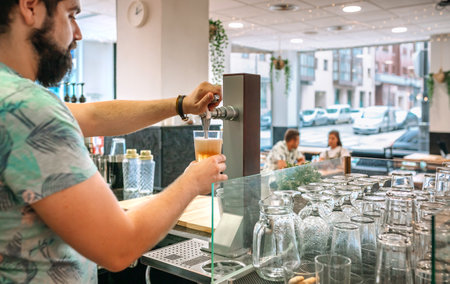 Bartender pouring draught beer from a tap in a restaurant, with customers sitting at a table in the background and glasses ready to be used. Image with copy space available on right.の写真素材