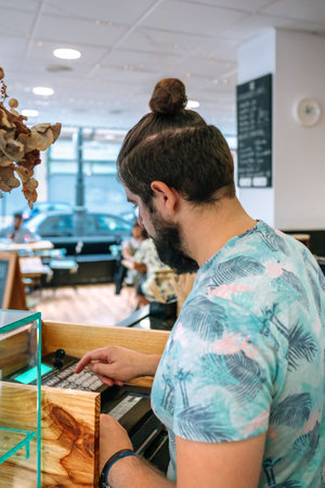 Young adult waiter with man bun and beard wearing stamped shirt using cash register in modern cafe with customers sitting at table in backgroundの写真素材