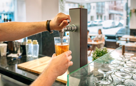 Close up of unrecognizable bartender hands pouring draught beer from a tap in a restaurant, with customers sitting at a table in the background and glasses ready to be usedの写真素材