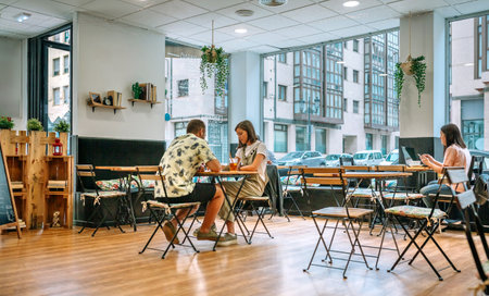 Young people sitting at tables in cafe with big windows, drinking juice and working on laptop, enjoying relaxing atmosphere during their leisure timeの写真素材