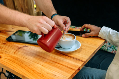 Unrecognizable barista preparing a frothy cappuccino, skillfully pouring creamy milk into a coffee cup on a rustic wooden table in a cozy cafe, while a customer waits nearby, engaged with a tabletの写真素材