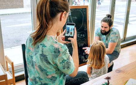 Father teaching to daughter on blackboard in their restaurant while mother takes pictures, capturing a moment of family and work balance. Restaurant owners family managing business and childcare.の写真素材